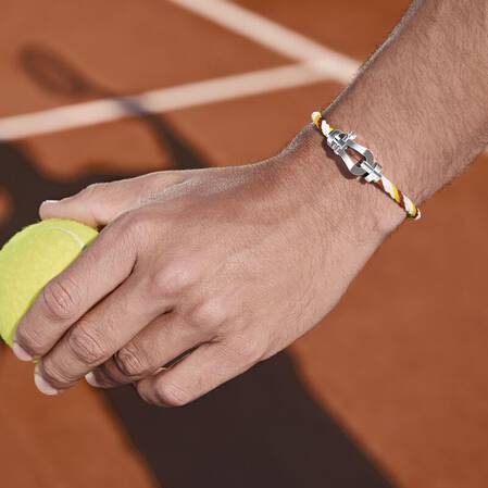 FRED x Roland-Garros : sur le court en terre battue, la main d’un joueur de tennis serre une balle jaune, mise en valeur par un bracelet tressé orné d’une boucle en or blanc étincelante Force 10.