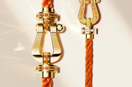 Close-up of two luxurious gold clasps adorned with yellow and orange gemstones, securing braided orange textile cords against a soft beige background.