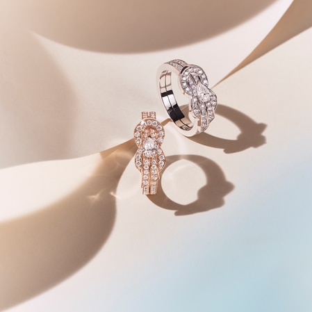 Two diamond knot-design rings, one in white gold and one in rose gold, rest on a softly lit surface casting distinct shadows.