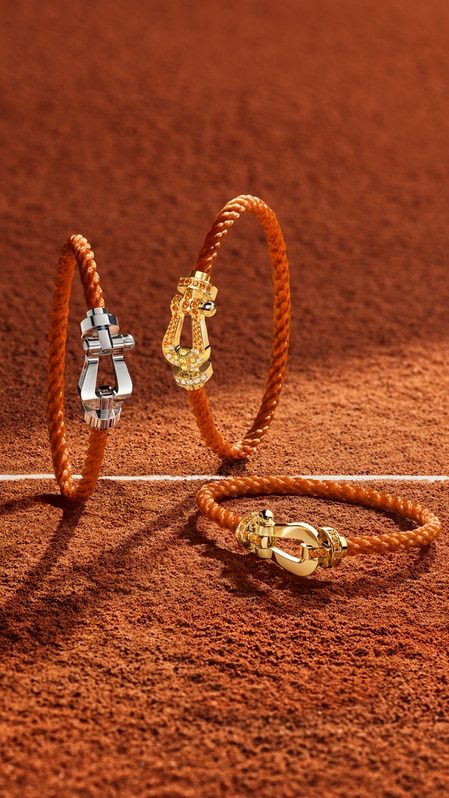 Trois bracelets &agrave; cordes de style &eacute;questre avec des fermoirs m&eacute;talliques, incluant de l'or blanc et de l'or jaune orn&eacute; de pierres pr&eacute;cieuses, reposant sur une surface textur&eacute;e de court de tennis en terre battue brun-rouge&acirc;tre, pr&egrave;s d'une ligne blanche.